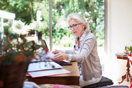 simsearch:614-08578614,k - Senior woman sitting at table, looking through paperwork, using laptop Photographie de stock - Premium Libres de Droits, Code: 649-09209551