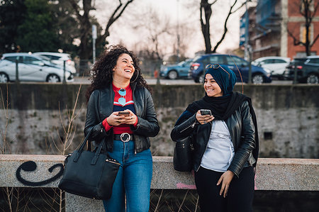 Young woman in hijab and her friend using smartphones and laughing by city canal Stock Photo - Premium Royalty-Free, Code: 649-09269087