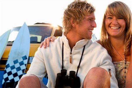 simsearch:649-01695983,k - Couple sitting on beach smiling with van and surfboards in background. Fotografie stock - Premium Royalty-Free, Codice: 649-01695947