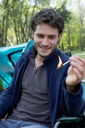 Man sitting at campsite holding a lit match smiling. Stock Photo - Premium Royalty-Free, Code: 649-01696214