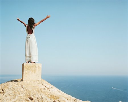 Woman standing on a large cement block with arms outstretched looking out over the sea. Stock Photo - Premium Royalty-Free, Code: 649-01696269