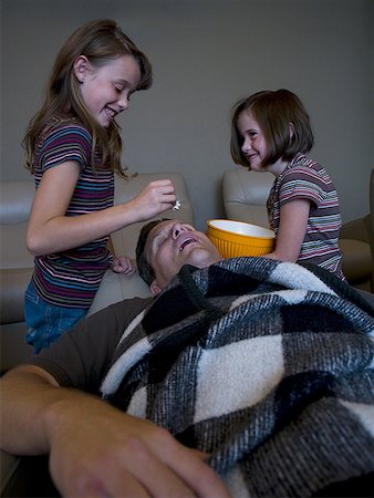 family watching tv together with popcorn - Daughters dropping popcorn into sleeping father's mouth Stock Photo - Premium Royalty-Free, Code: 640-01358340