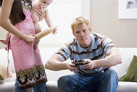 Man on sofa with video game controller and woman in apron with rolling pin holding baby Stock Photo - Premium Royalty-Free, Code: 640-01458917