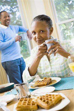 Portrait of a senior woman sitting at the breakfast table with a senior man standing behind her Stock Photo - Premium Royalty-Free, Code: 640-02764586