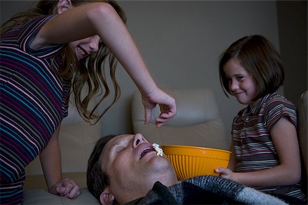 family watching tv together with popcorn - Daughters dropping popcorn into sleeping father's mouth Stock Photo - Premium Royalty-Free, Code: 640-02769033