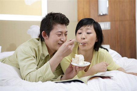 photo of a woman feeding her husband food - Husband feeding wife with fork while lying down on bed Stock Photo - Premium Royalty-Free, Code: 642-01735516
