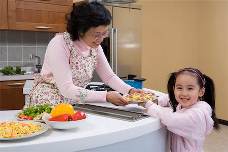 Senior woman with granddaughter holding plate in kitchen and smiling Stock Photo - Premium Royalty-Free, Code: 642-01736637