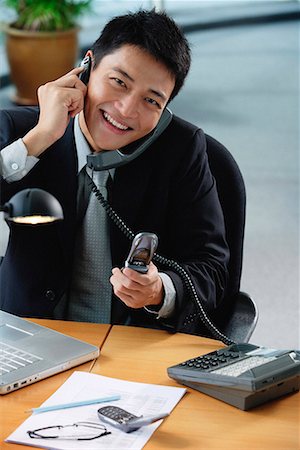 simsearch:656-01766255,k - A man smiles at the camera as he sits at his desk which has lots of phones on it Stock Photo - Premium Royalty-Free, Code: 656-01766174