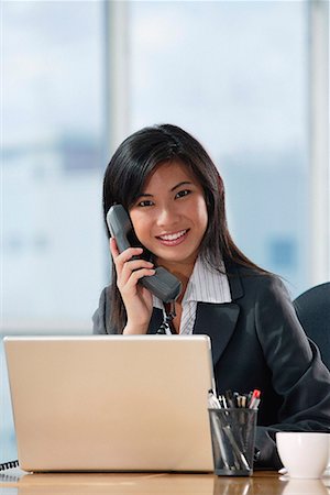 simsearch:656-01766255,k - A woman looks at the camera and talks on the phone as she sits at her desk Stock Photo - Premium Royalty-Free, Code: 656-01766376