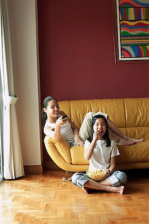Two girls sitting in living room, one holding remote control, the other eating popcorn Stock Photo - Premium Royalty-Free, Code: 656-01768242