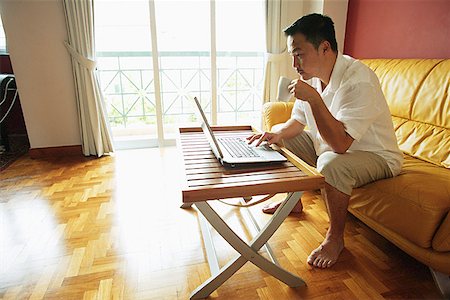 simsearch:656-03519571,k - Man sitting in living room, using laptop, drinking from mug Foto de stock - Sin royalties Premium, Código: 656-01768273