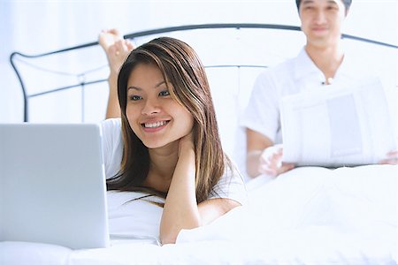 simsearch:656-01773393,k - Couple sur le lit, femme à l'aide de man portable derrière elle, lire le journal Photographie de stock - Premium Libres de Droits, Code: 656-01768929