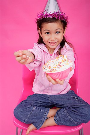Girl with party hat, sitting on chair, holding bowl of cake towards camera Stock Photo - Premium Royalty-Free, Code: 656-01772772