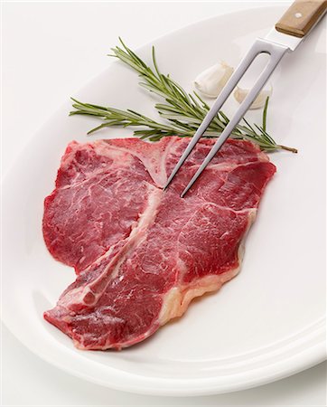 Raw T-bone steak with carving fork, rosemary and garlic Fotografie stock - Premium Royalty-Free, Codice: 659-06187555