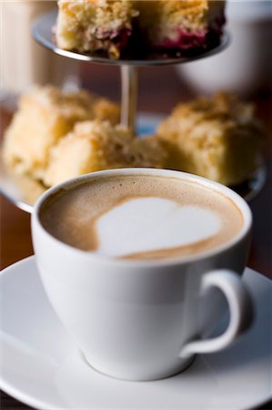 A cappuccino decorated with a heat with a cake stand in the background Foto de stock - Sin royalties Premium, Código: 659-06306412