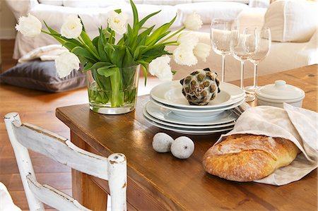 A bunch of white tulips in a glass vase next to a stack of plates, some wine glasses, decorative figs and a decorative artichoke Foto de stock - Sin royalties Premium, Código: 659-07027652