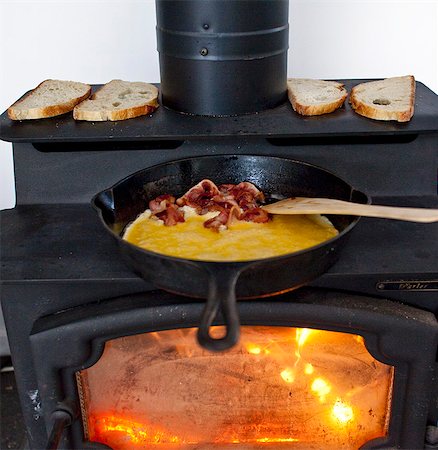 queimado - Eggs and Bacon Cooking in a Cast Iron Skillet on a Wood Burning Stove; Bread Toasting on the Stove Foto de stock - Royalty Free Premium, Número: 659-07027865