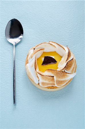 An individual lemon meringue tart and a spoon on a blue surface Photographie de stock - Premium Libres de Droits, Code: 659-07068862