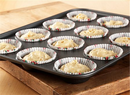 Uncooked cupcake mixture in cupcake cases and a cupcake tin sitting on a wooden board all on a wooden table Stock Photo - Premium Royalty-Free, Code: 659-08897074
