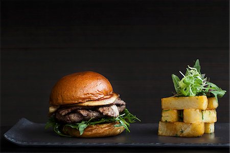 simsearch:659-07069745,k - A juicy burger with bacon fois gras and lettuce in a bun served with a stack of chunky square cut chips on a black background Photographie de stock - Premium Libres de Droits, Code: 659-08897343