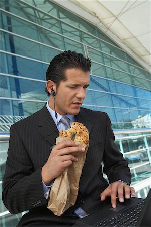 people eating bagels - Businessman eating bagel while using laptop with earpiece in outside office building Stock Photo - Premium Royalty-Free, Code: 694-03327802
