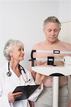 female doctor with male patient - Senior medical practitioner stands with clip-board and client on measuring scales Stock Photo - Premium Royalty-Free, Code: 694-03332847