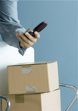 delivery mobile - Man's hand holding cell phone, cardboard boxes piled on chair Stock Photo - Premium Royalty-Free, Code: 695-05764144