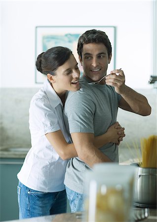 In kitchen, woman holding man around waist as man reaches back with spoon for woman to taste Stock Photo - Premium Royalty-Free, Code: 695-05764209