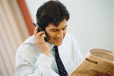 Businessman using phone and holding up financial section of newspaper Stock Photo - Premium Royalty-Free, Code: 695-05764532