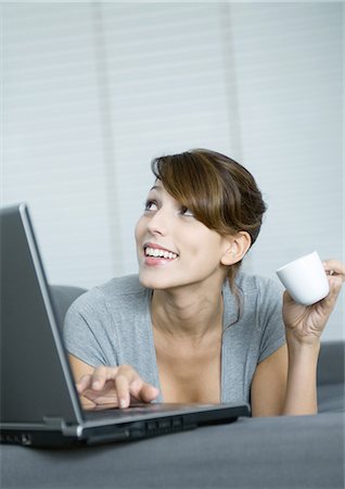 simsearch:649-08144347,k - Young woman on couch, using laptop and holding coffee cup, looking up Foto de stock - Royalty Free Premium, Número: 695-05764811