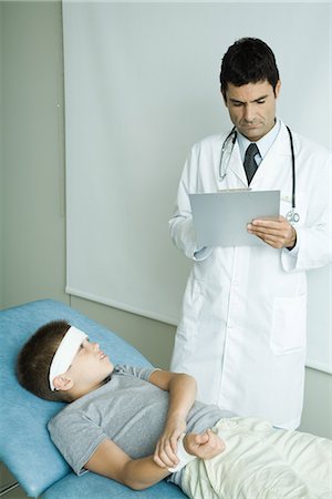 Boy lying on examination table with bandage on forehead, holding arm, doctor writing on clipboard Stock Photo - Premium Royalty-Free, Code: 695-05765481