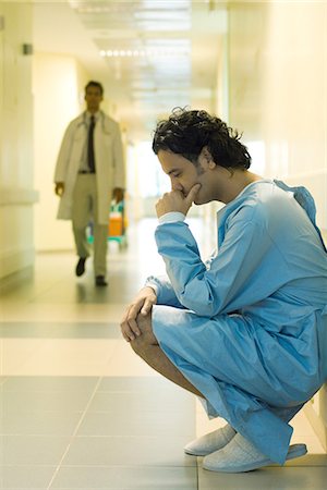 Männliche Patienten im Krankenhaus Korridor hocken, halten Kopf, Blick nach unten, Arzt im Hintergrund laufen Stockbilder - Premium RF Lizenzfrei, Bildnummer: 695-05766637