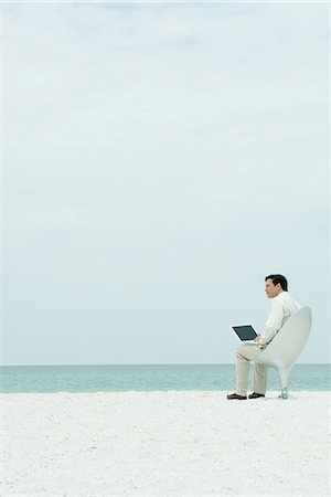 Businessman sitting in chair on beach, using laptop computer, looking away Stock Photo - Premium Royalty-Free, Code: 695-05767050