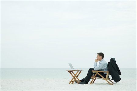 Businessman sitting on beach, looking at laptop computer, hand under chin, full length Stock Photo - Premium Royalty-Free, Code: 695-05767069