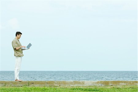 easy - Man standing on low wall using laptop, body of water in background, full length Stock Photo - Premium Royalty-Free, Code: 695-05767499