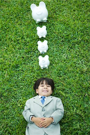 front view of a pig - Little boy in full suit lying on grass, line of piggy banks above head Stock Photo - Premium Royalty-Free, Code: 695-05767535