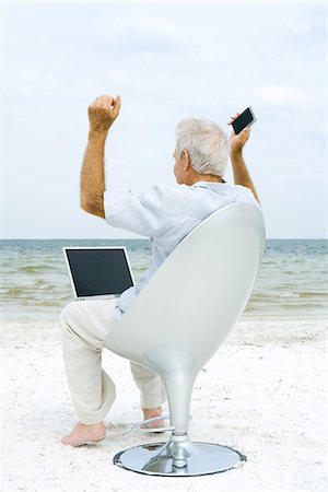 Senior man using laptop and cell phone on beach, raising arms Stock Photo - Premium Royalty-Free, Code: 695-05767531
