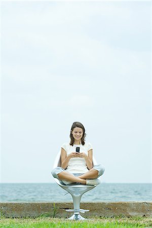 Teenage girl sitting in chair by the sea, looking down at cell phone, smiling Stock Photo - Premium Royalty-Free, Code: 695-05767582