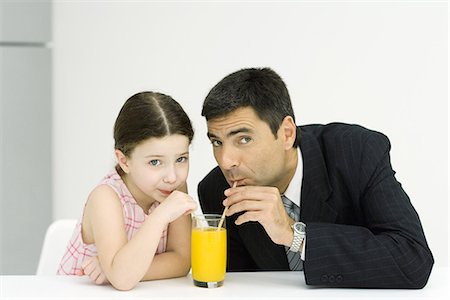 Little girl and father drinking from glass of juice with straws, looking at camera Stock Photo - Premium Royalty-Free, Code: 695-05767944