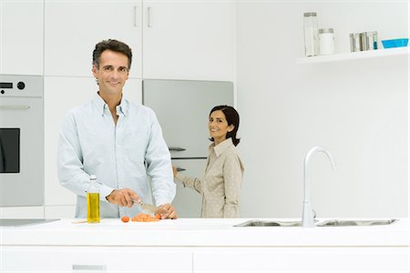 Man and woman in kitchen, man chopping tomatoes while woman goes toward refrigerator, both smiling at camera Stock Photo - Premium Royalty-Free, Code: 695-05767946