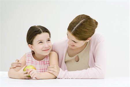 Woman sitting at table with arm around daughter's shoulder, smiling, girl holding apple Stock Photo - Premium Royalty-Free, Code: 695-05768177