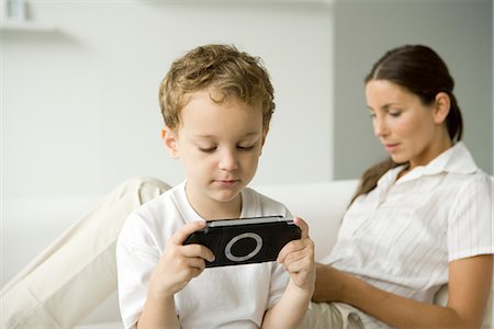 simsearch:6108-07969509,k - Little boy playing handheld video game, his mother sitting on sofa behind him Stock Photo - Premium Royalty-Free, Code: 695-05768281