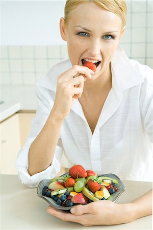 simsearch:695-03378760,k - Woman holding bowl of fruit salad, eating strawberry, looking at camera Foto de stock - Sin royalties Premium, Código: 695-05768338