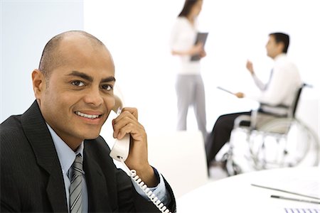 Businessman using phone, smiling at camera, colleagues in background Stock Photo - Premium Royalty-Free, Code: 695-05768371