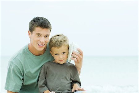 Father holding seashell up to son's ear, both smiling at camera Stock Photo - Premium Royalty-Free, Code: 695-05768589