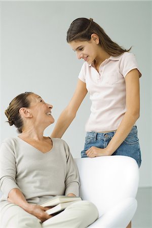 simsearch:400-05697219,k - Grandmother sitting in chair, holding book, smiling up at teen granddaughter standing nearby Stock Photo - Premium Royalty-Free, Code: 695-05768693