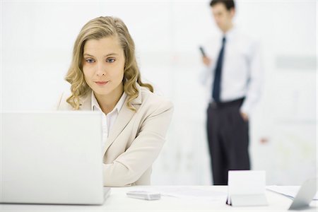 raised eyebrow businessman - Professional woman using laptop computer at desk, man standing in background Stock Photo - Premium Royalty-Free, Code: 695-05768874