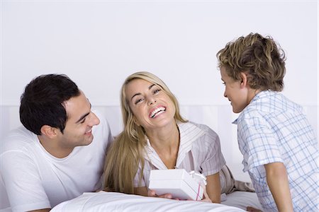 simsearch:695-05769105,k - Family lying in bed together, woman holding gift and smiling at camera Stock Photo - Premium Royalty-Free, Code: 695-05769103