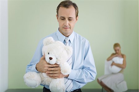 New father holding teddy bear, looking down, wife holding baby in background Stock Photo - Premium Royalty-Free, Code: 695-05769114