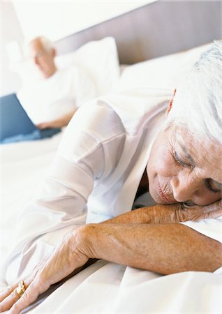 Senior woman sleeping on bed, man using laptop in background Stock Photo - Premium Royalty-Free, Code: 695-05774051